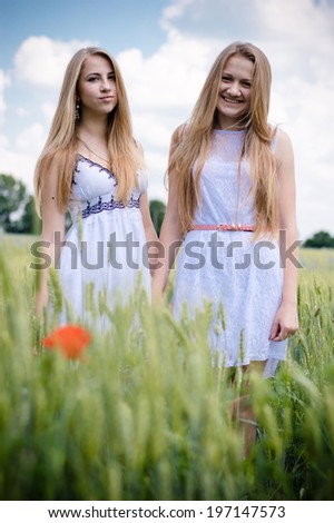 2 young women happy smiling blond girl friends walking in green field & looking at camera on summer outdoors blue sky copy space background portrait image