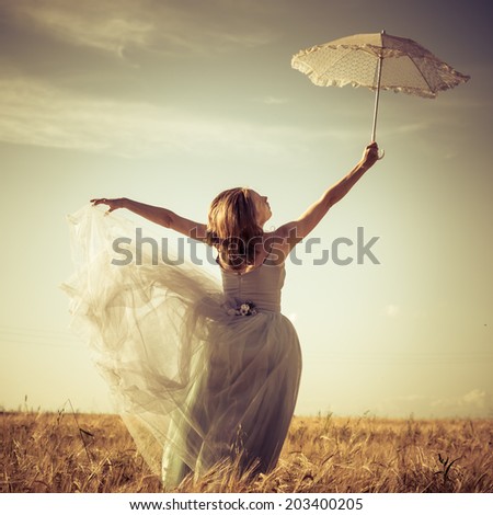 romantic summer outdoors fairy: beautiful blond young woman having fun wearing long blue ball dress and holding white lace umbrella leaning up on wheat field and blue sky copy space background