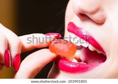closeup image on fingers with red color nails holding candy at seductive charming beautiful red lips & excellent dental whitening care teeth