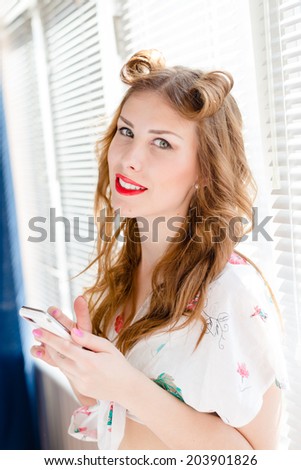 having fun reading message on mobile cell phone beautiful young pinup lady happy smile & looking at camera on sun lighted window blinds copy space background closeup portrait picture