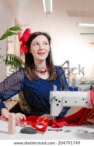 Funny young pinup pretty girl having fun with sewing machine and measuring tape happy smile & looking up on white or light workshop wall copy space background portrait image