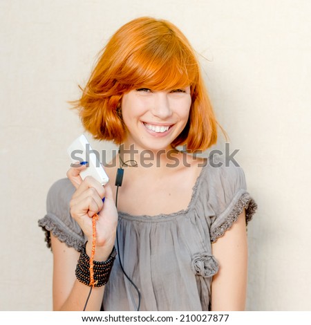 portrait of happy smiling beautiful young woman with red hair having fun holding mobile digital device or mobile phone & looking at camera on light copy space background wall