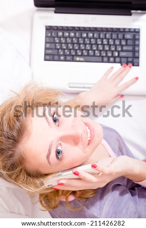 top view on attractive young business woman in bed in pajamas talking on the mobile smart phone and working on laptop happy smiling & looking at camera closeup portrait