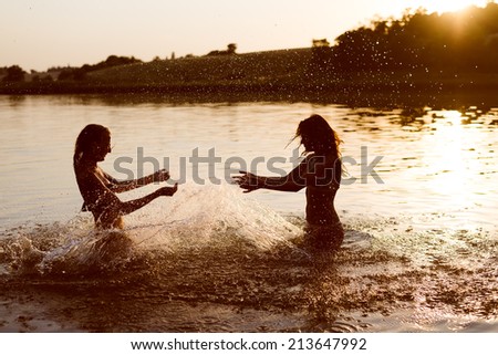 summer fun: filtered image of 2 beautiful young woman or teenage girls best friends having fun and splashing water in river or lake at sunset on sunny outdoors copy space background