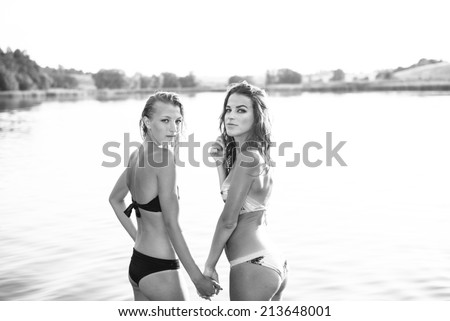 black and white image of 2 attractive young women or teenage girls best friends in bikini holding hands and looking over shoulder in camera on summer river or lake outdoors copy space background