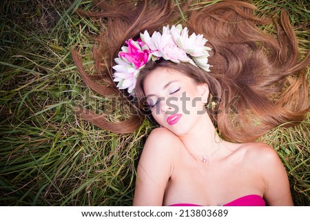 sleeping beauty: filtered image of beautiful brunette young woman in lotus flower crown and pink makeup lying gracefully on green grass outdoors copy space background