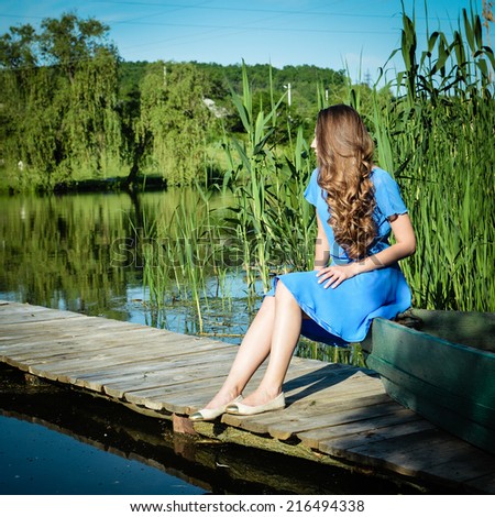 portrait of romantic young brunette pretty lady having fun relaxing sitting in blue dress on river or lake pier dreaming on sunny day copy space background