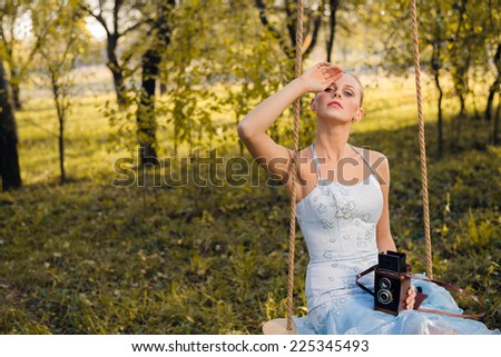 Beautiful young woman in prom or cocktail dress sitting on swings with retro photo camera on green summer or autumn outdoors copyspace background