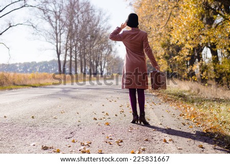 Young lady in retro hat and overcoat with vintage suitcase waiting on empty autumn road