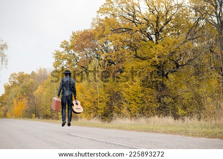 Young man musician in retro hat and leather jacket with vintage suitcase and guitar jumping or flying on empty autumn road copy space background