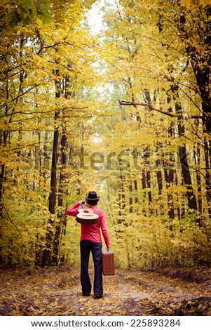 Young man musician in retro hat and leather pants with vintage suitcase and guitar wondering in yellow autumn forest copy space background