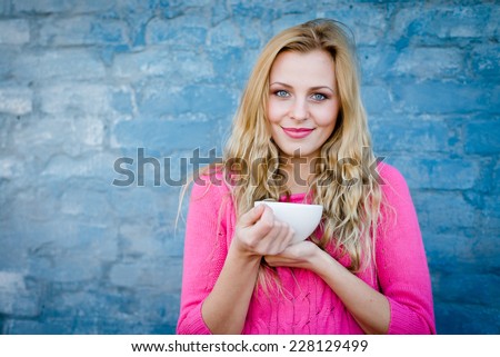 Happy young blond woman enjoying hot tea or coffee and smiling over blue brick wall copy space background