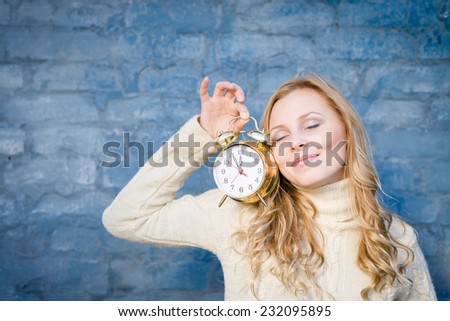 Pretty blond teenage girl with golden alarm clock smiling at blue brick wall background copy space image