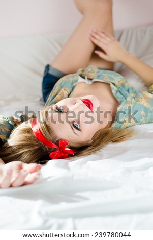 portrait of young beautiful blonde woman in jeans shorts floral shirt lying on back white bed putting your feet on wall & looking at camera