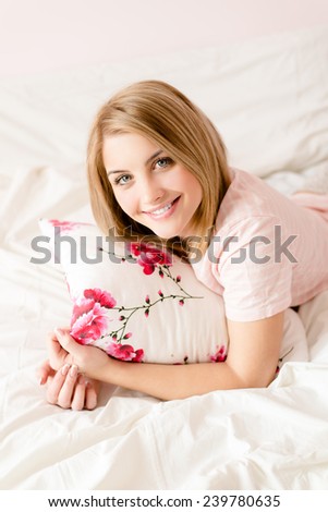 portrait of attractive happy young blond woman in bed with floral pillow in hand  & looking at camera