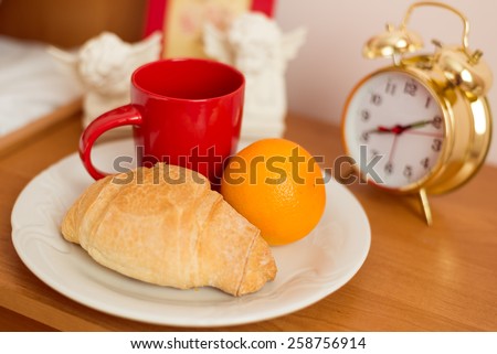 Picture of croissant, orange with red cup on white plate and alarm clock