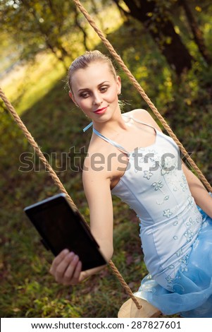 Beautiful young woman in prom dress sitting on swing and making selfie by tablet pc on green summer outdoors