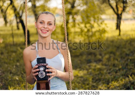 Beautiful young woman in prom dress sitting on swing with retro photo camera on green summer outdoors