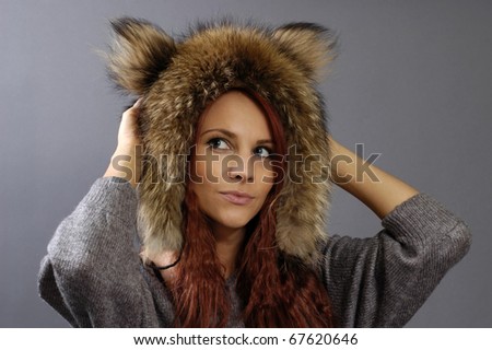 The young beautiful woman in a winter cap from fur of a raccoon on a gray background