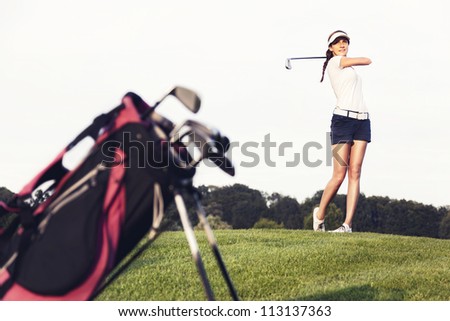 Girl golf player hitting ball on golf course with golf bag in foreground.
