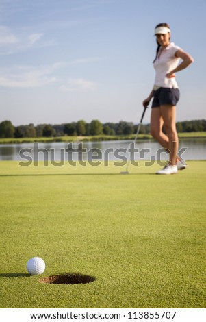 Relaxed woman golf player looking at ball that stopped near cup after putting on green. Focus on ball and hole.