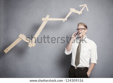 Concept: Successful business. Confident young businessman on the mobile-phone in front of rising business graph, isolated on grey background.