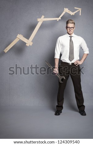Concept: Building your own successful career or business. Young confident businessman holding  wrench in front of business graph with positive trend, isolated on grey background.