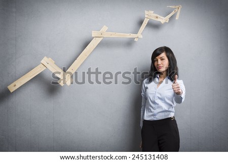 Concept: Successful career or business. Young confident businesswoman holding thumb up in front of ascending business graph, isolated on grey background.