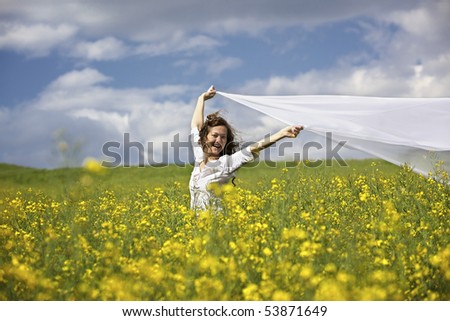 Young happy woman standing in yellow rapeseed field holding a white long piece of cloth in the wind.