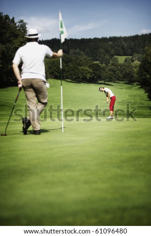 Attractive female golf player putting on green with second male player in the foreground holding the flag.