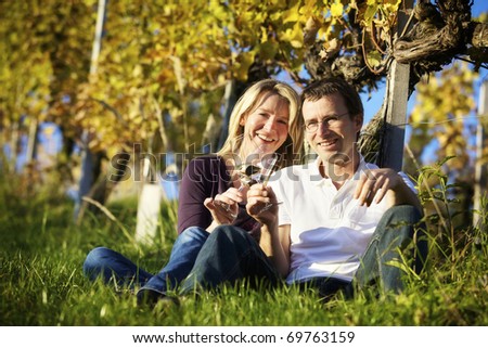 Cheers! Friendly woman and man (couple) sitting in vineyard holding glasses for wine drinking.