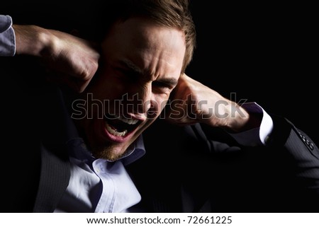 Low-key portrait of desperate businessman in dark suit screaming and holding both fists at head expressing strong despair, isolated on black background.