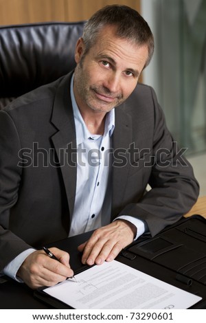 Smiling businessman in dark suit and blue shirt sitting at office desk and signing a contract, looking up.