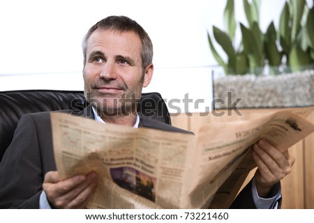 Smiling business person in dark suit sitting in office chair reading newspaper and looking up.