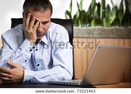 Senior business person in blue shirt sitting at office desk looking at laptop and being frustrated about computer or software crash.