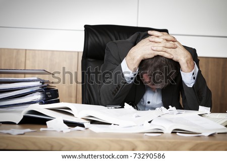 Worried businessman in dark suit sitting at office desk full with books and papers being overloaded with work.