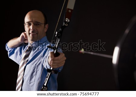 Handsome smiling businessman in blue short and tie hitting the target with bow and arrow, isolated on black background.