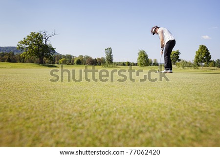 Active senior female golf player focusing for putting golf ball on green on beautiful golf course with blue sky in background.