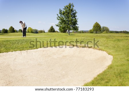 Active senior female golf player putting golf ball on green on beautiful golf course with sand bunker in foreground.