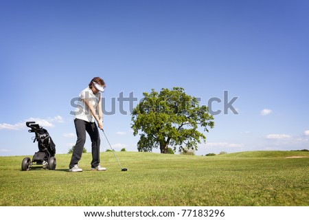Active senior female golf player with golf bag concentrating to hit the ball with driver on fairway on beautiful golf course with blue sky in background.