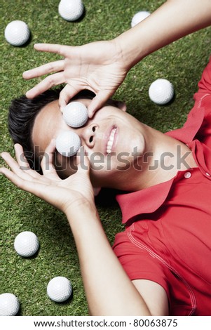 Young smiling male golf player in red shirt lying on green next to several golf balls and covering his eyes with two golf balls.