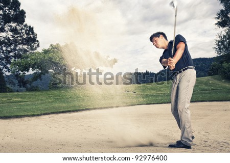 Male golfer in blue shirt and grey pants hitting golf ball out of a sand trap with sand wedge and sand caught in motion.