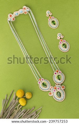 Soutache bijouterie set colorful earrings and necklaces with green purple cyan and pink crystals on green background with grass stems and yellow flowers