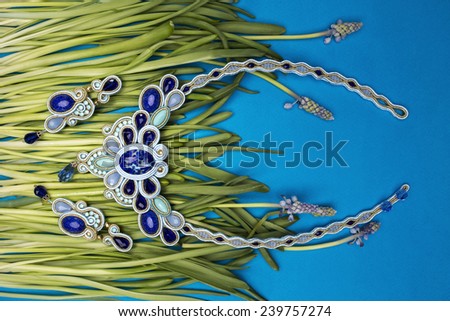 Soutache bijouterie set with blue earrings and necklaces with blue stones and light-blue and cyan crystals on the cyan background of grass stems with purple flowers