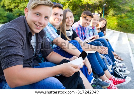 group of young students with books and gadgets sit on the steps in the park