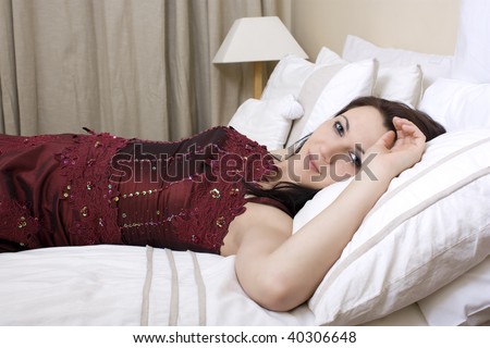 A beautiful woman laying on a bed with white duvet and pillows, wearing a dark red evening gown/dress.