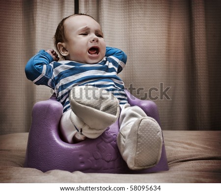 Close up studio shot of a baby boy sitting on a rubber baby support chair on the bed screaming out loud.