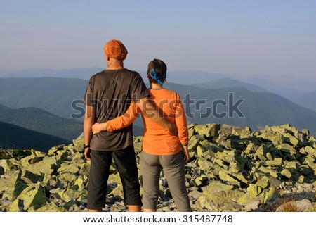 Rear view, couple hikers looking into the distance, is standing on the top of mountain. Background is mountains and blue sky.