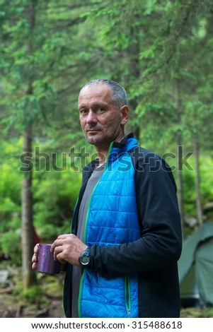 Hiker  is resting with cup of tea on the background coniferous forest. A  serious man with cap of tea.