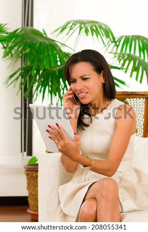 Beautiful young elegant woman holding digital tablet and talking on cell phone looking confused